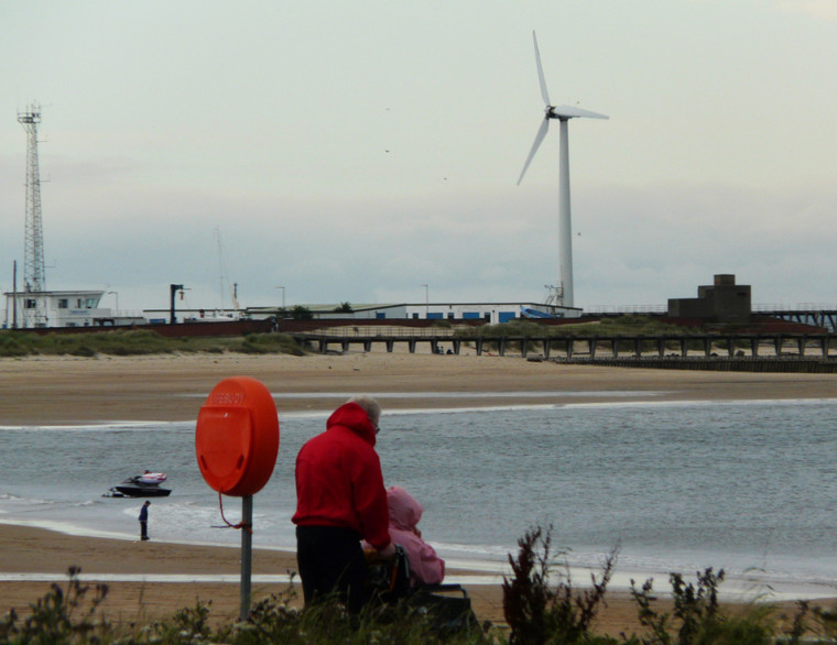 A person pushing another person in a wheelchair. They are by a beach, with wind turbines in the background.