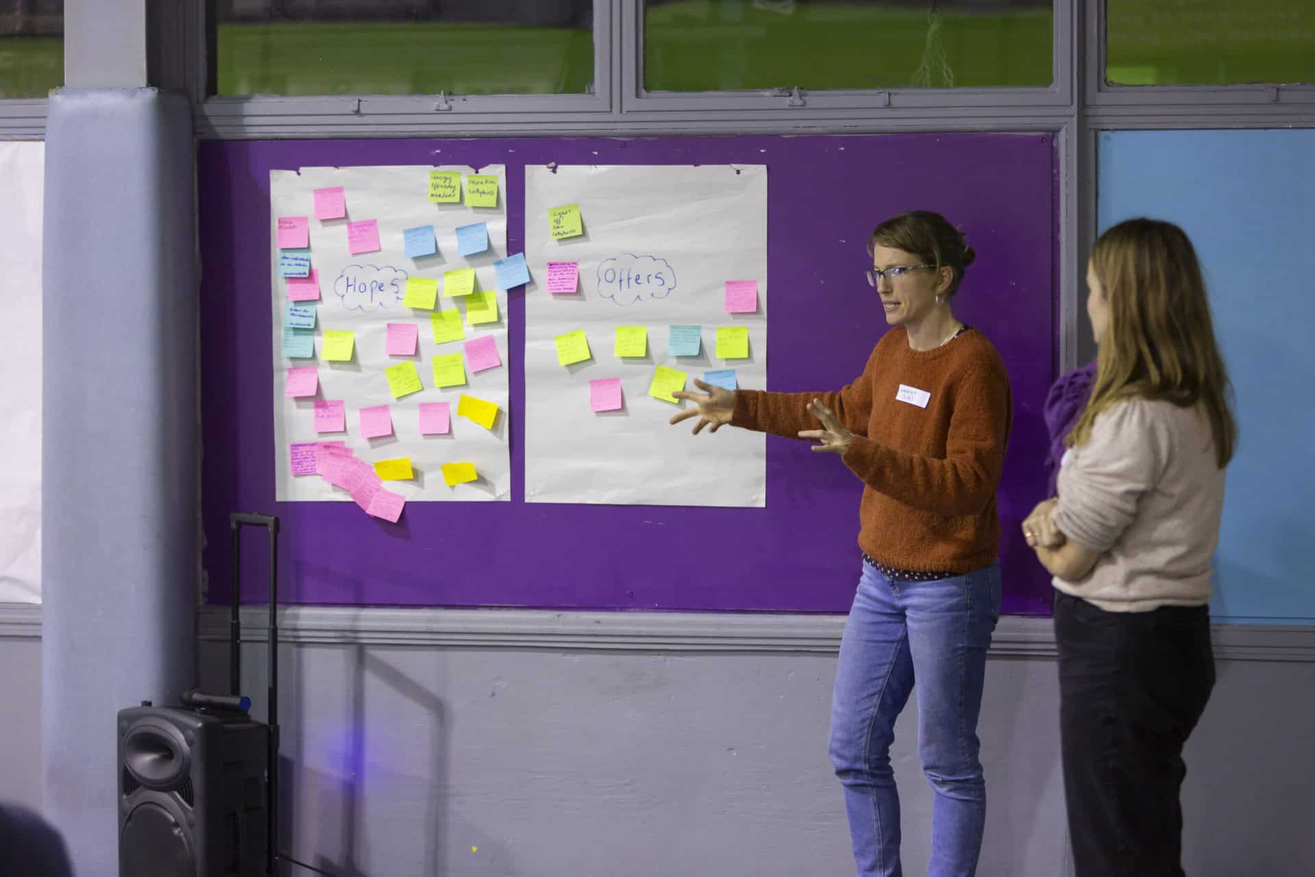 Two women stand in front of a wall with two pieces of paper pinned up. The pieces of paper are labelled 'hopes' and 'offered' and are covered with brighly coloured post-its.