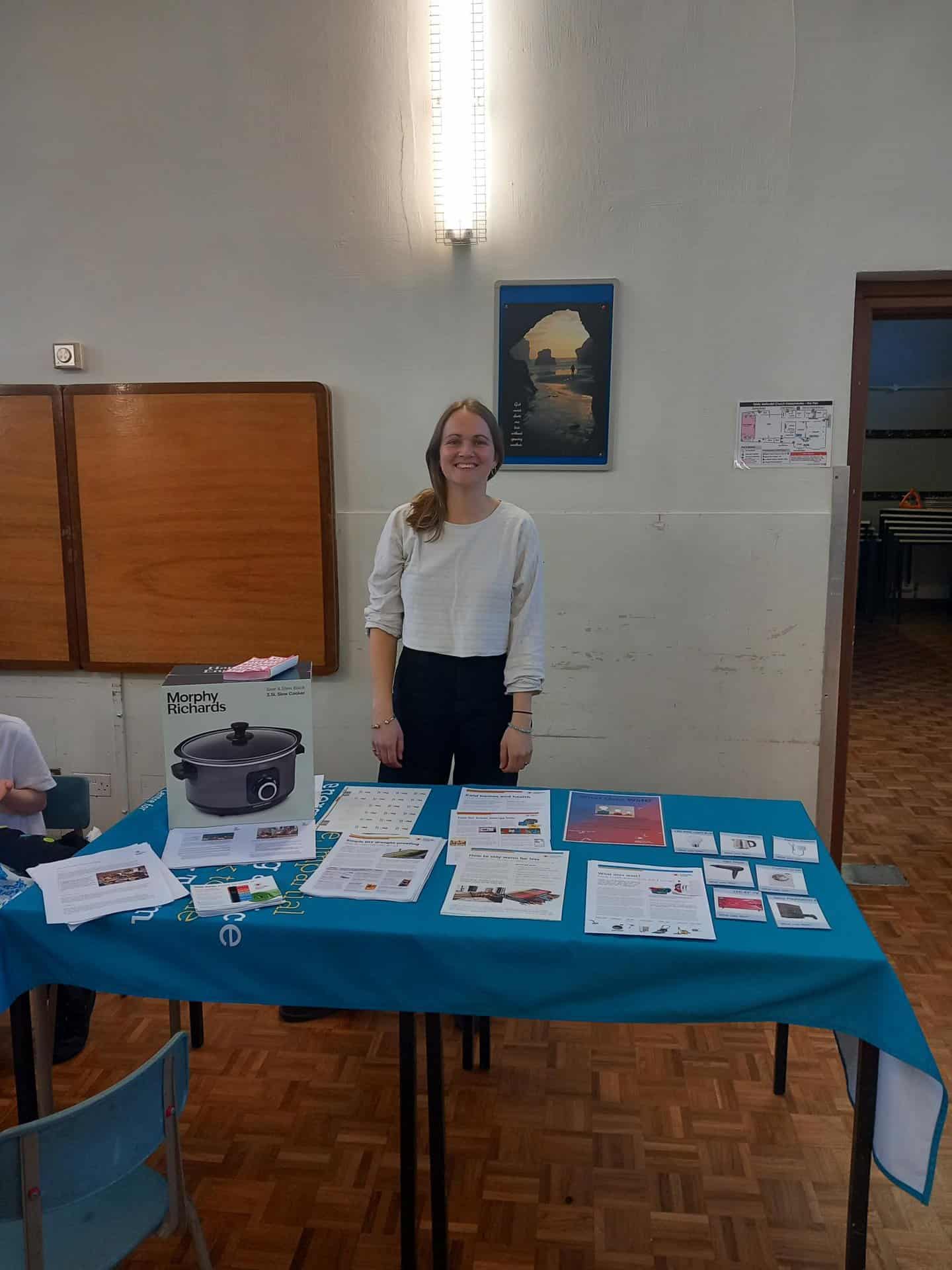 A smiling woman stands behind a table with a slow cooker and many pieces of paper on it.