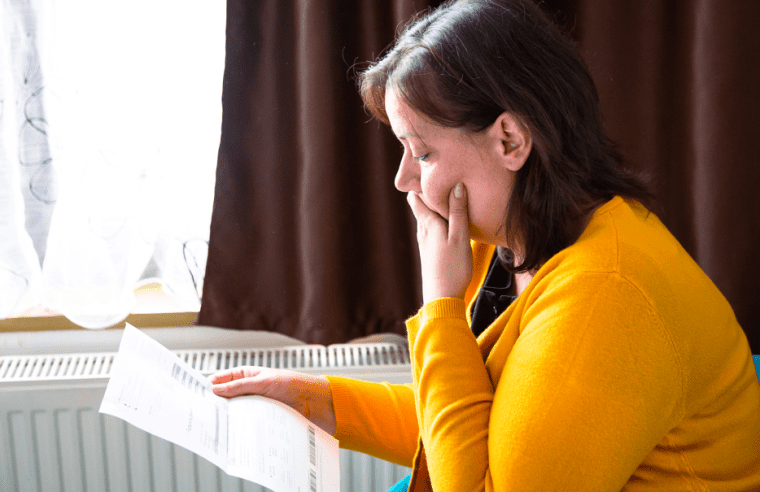 A woman sat on a sofa in a yellow jumper looks concerned while reading her latest energy bill
