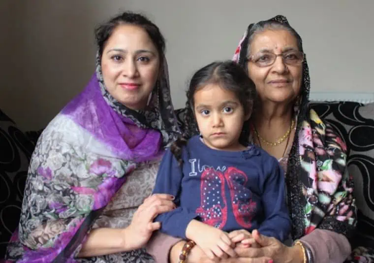 A South Asian family of mother, grandmother and daughter sit on a sofa smiling at the camera