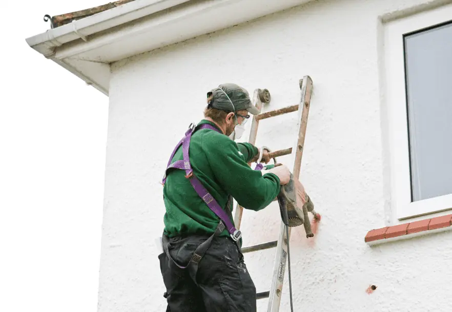 A worker on a ladder drilling holes in a cavity wall that will be filled with insulation