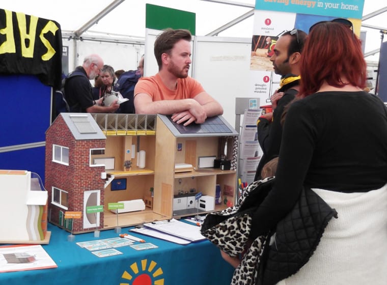 An event stand with a model of a wooden house on top. An energy advisor and community members stand around it talking about energy.