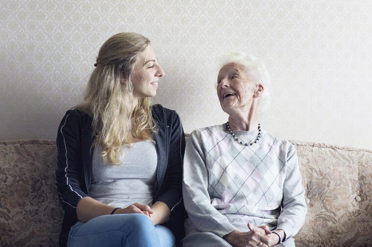 A young woman and an elderly woman sit next to each other on a sofa smiling at each other