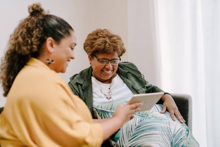 Two women sit on a sofa, one is showing the other something on an iPad. Both are smiling.