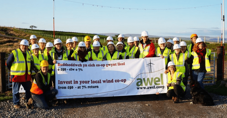 A group of people in high-vis jackets and white hard hats stand in a field holding a large sign saying 'Invest in your local wind co-op'