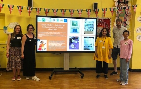 Three teachers and two trainers stand next to a presentation board during an event to highlight energy use