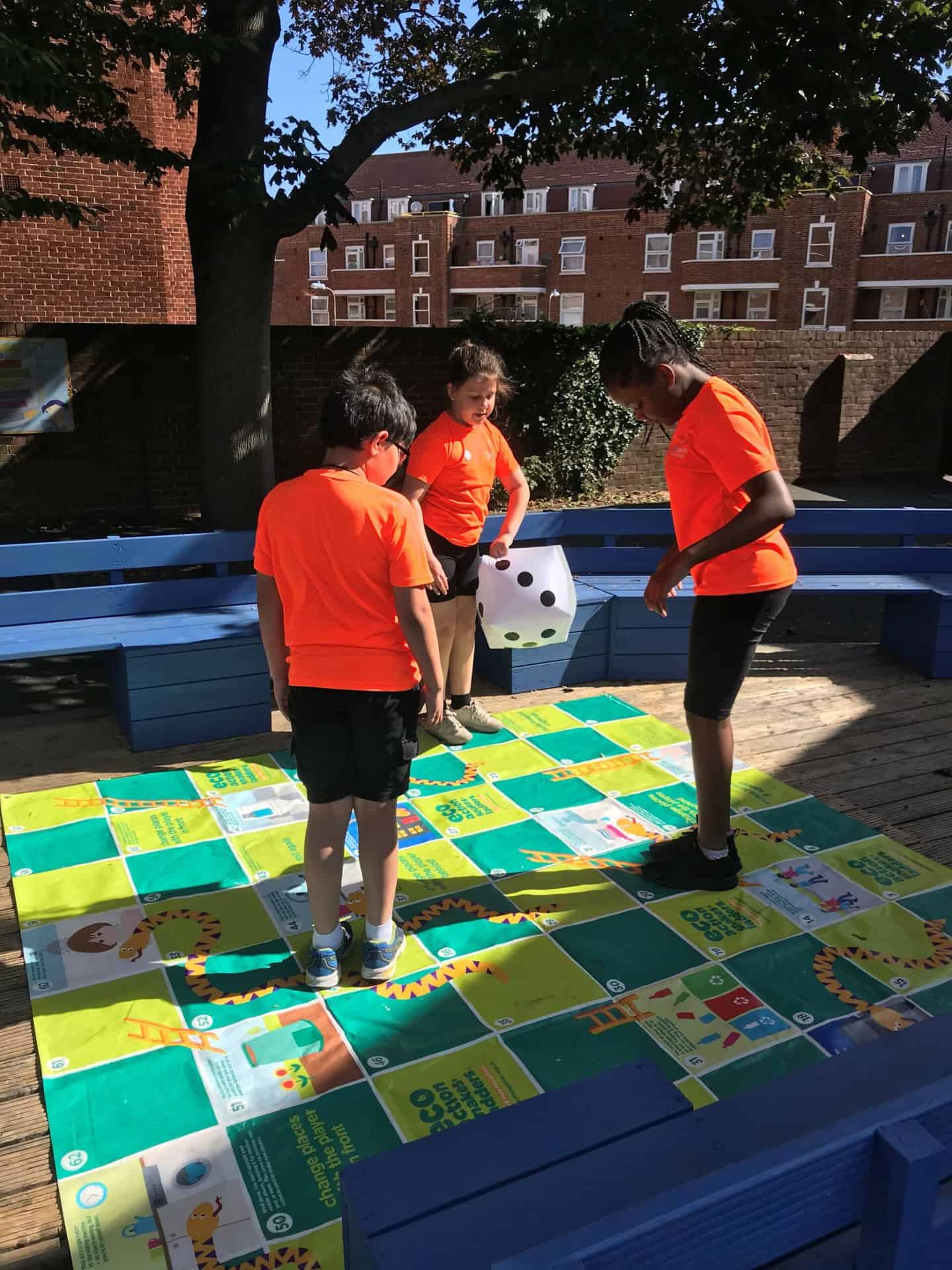Three young children play a game outside using a snakes and ladders board and large dice
