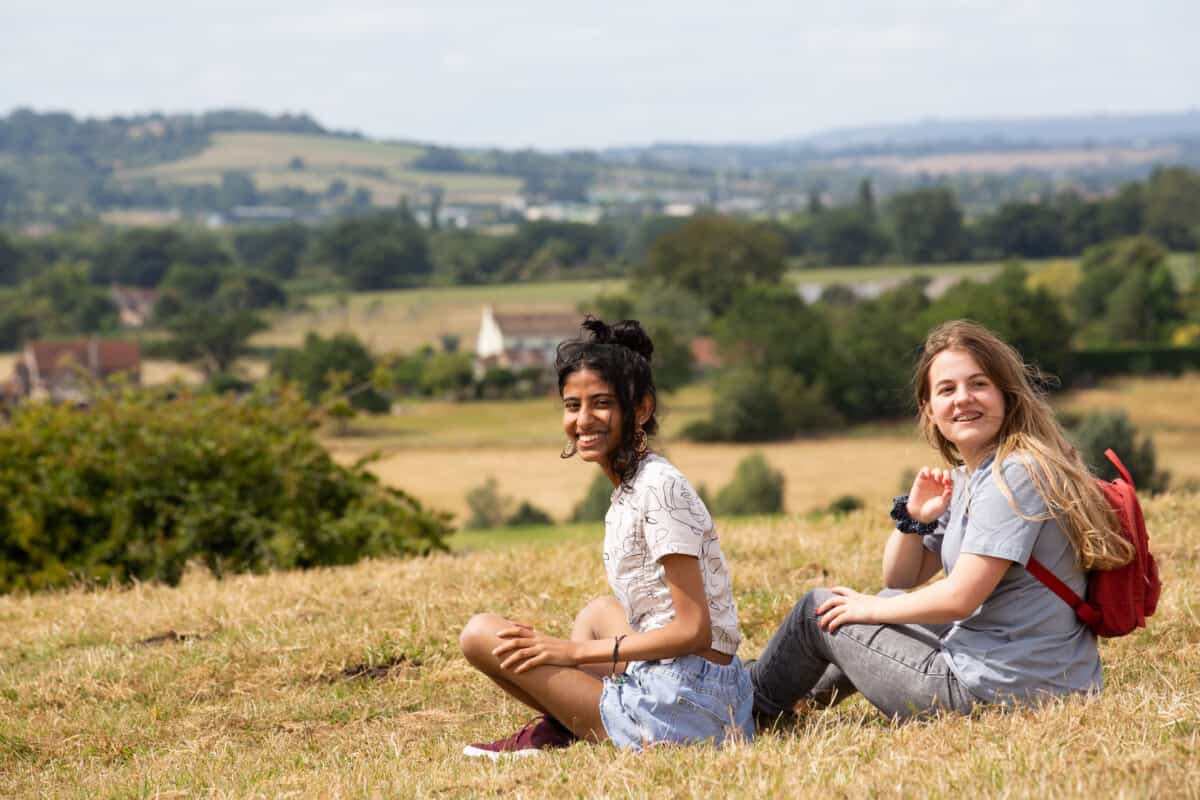 Two teenagers sit on a grassy bank. They are smiling towards the camera.