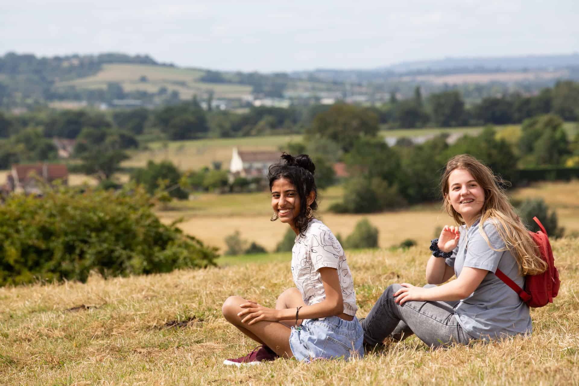 Two teenagers sit on a grassy bank. They are smiling towards the camera. 