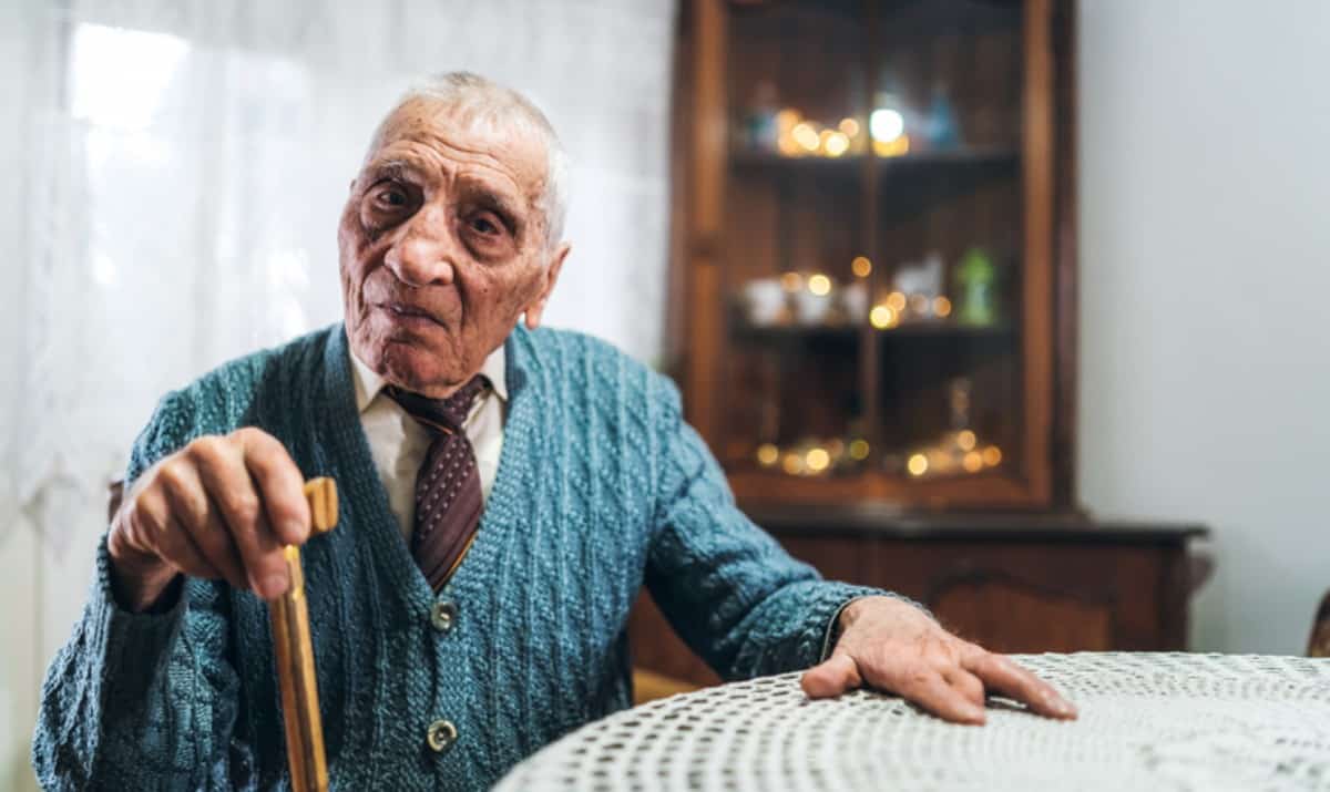 An older man sits at a table. He has a walking stick and is looking directly at the camera, deep in thought.