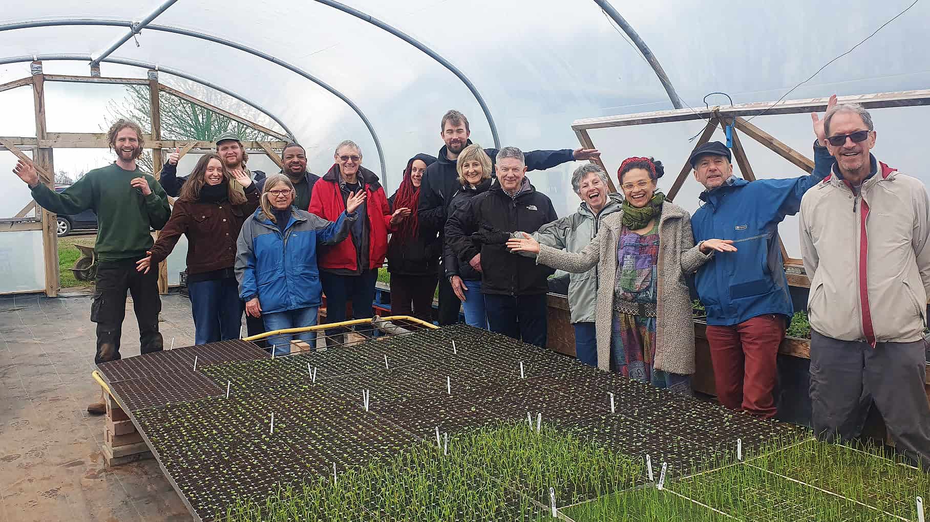 A group of people of all ages stand around a table covered in seedlings in a polytunnel and smile and wave at the camera.
