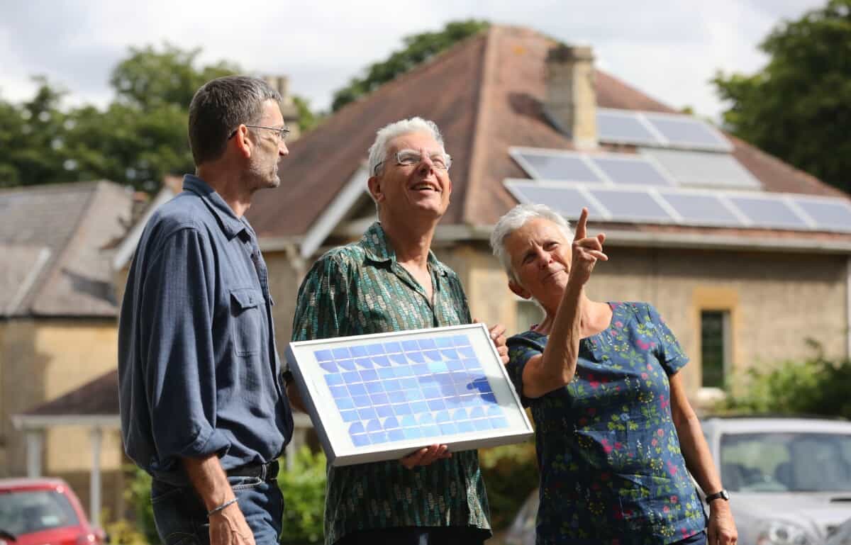 A group of three people - members of a community energy group - look at a house with solar panels on the roof. More solar panels can be seen on the house behind them.