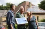 A group of three people - members of a community energy group - look at a house with solar panels on the roof. More solar panels can be seen on the house behind them.