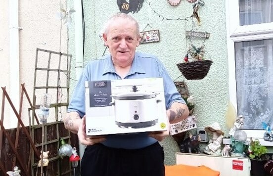 An elderly man, named Derek, stands at the front of his house holding a boxed slow cooker