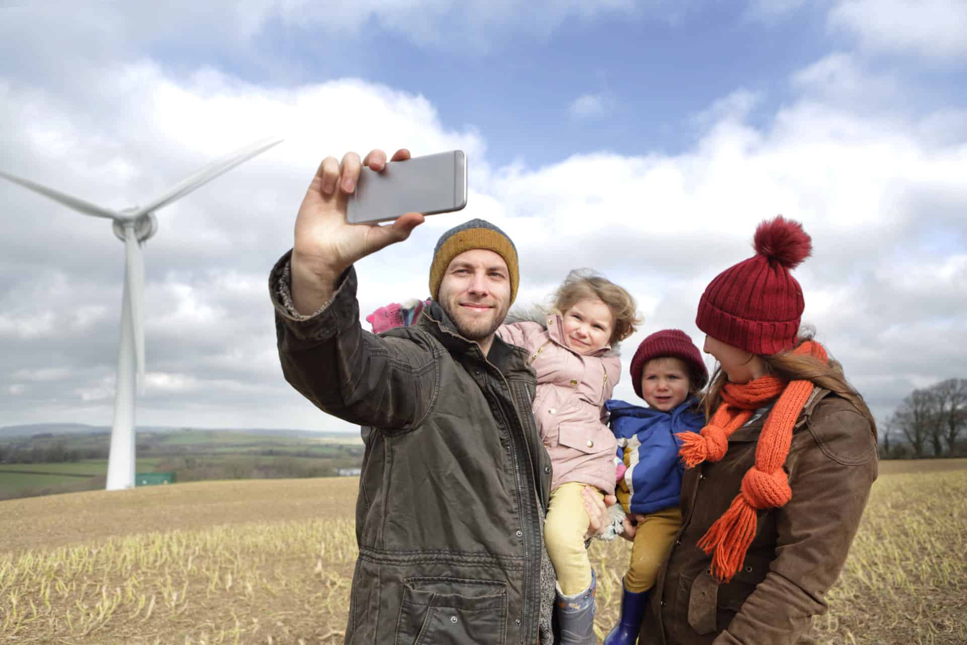 A family with very young children take a selfie in front of a wind turbine