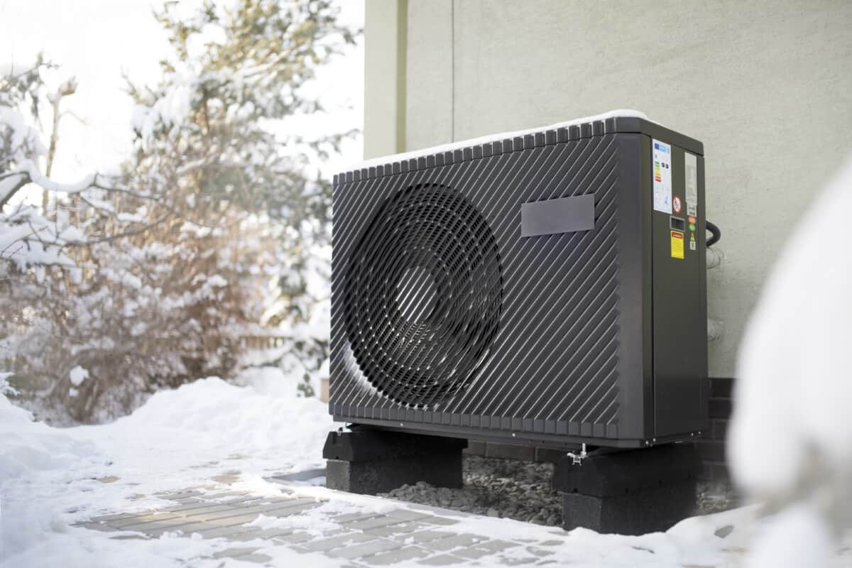 An air source heat pump standing beside a house with snow on the ground.