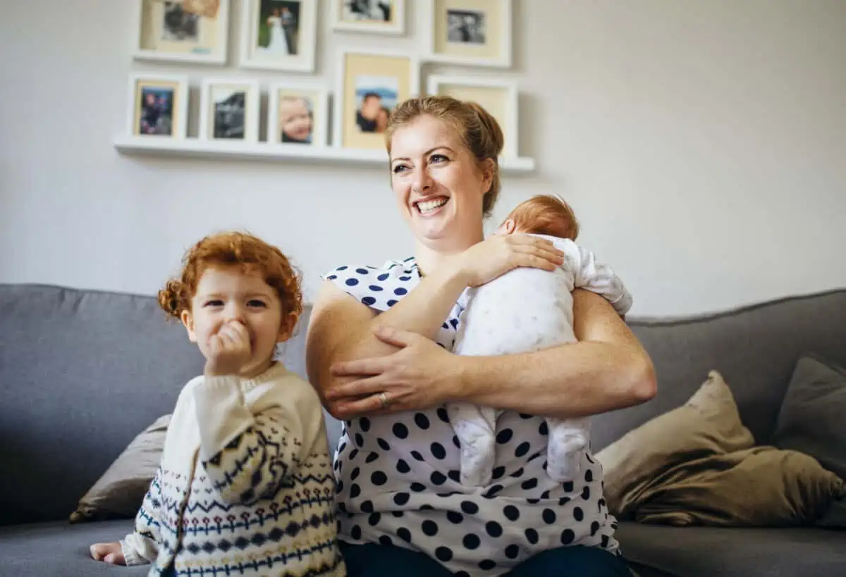 Mother with her two children in the living room of their home. The mother is holding a baby and the other little girl is laughing and looking at the camera.