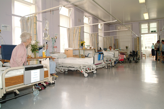 A hospital ward. A patient sits on their bed looking away from the camera.