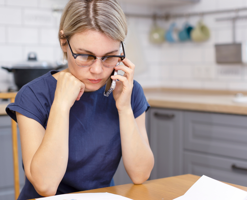 A worried-looking woman sits at her kitchen table looking at bills.