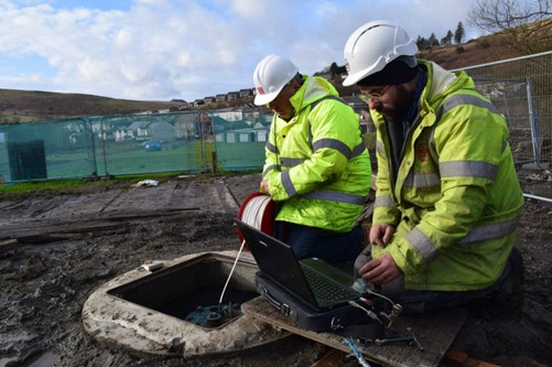 Two men in high-vis coats and hard hats kneeling next to an open manhole.