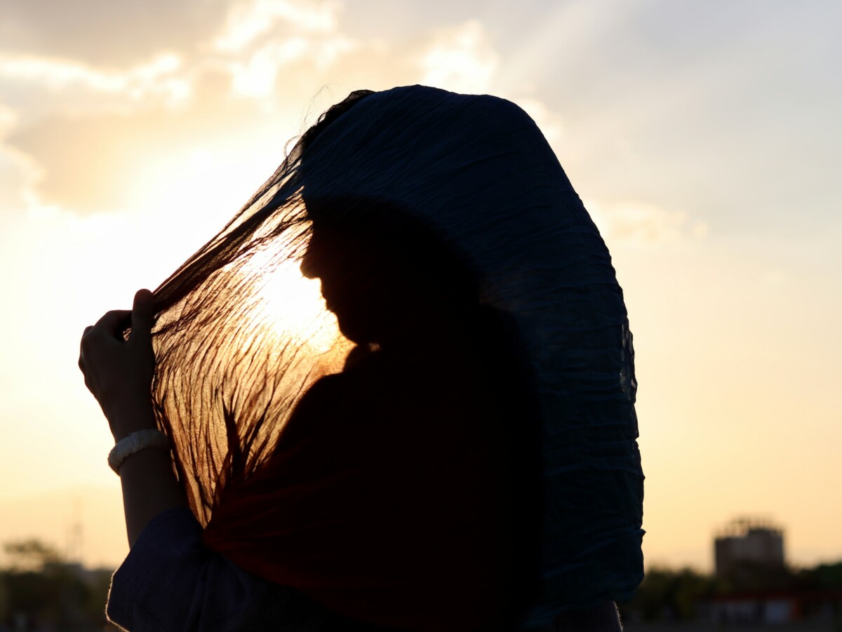 A woman wears a hijab and holds part of it over the sun. Her silhouette is outlined and we see her shadow.