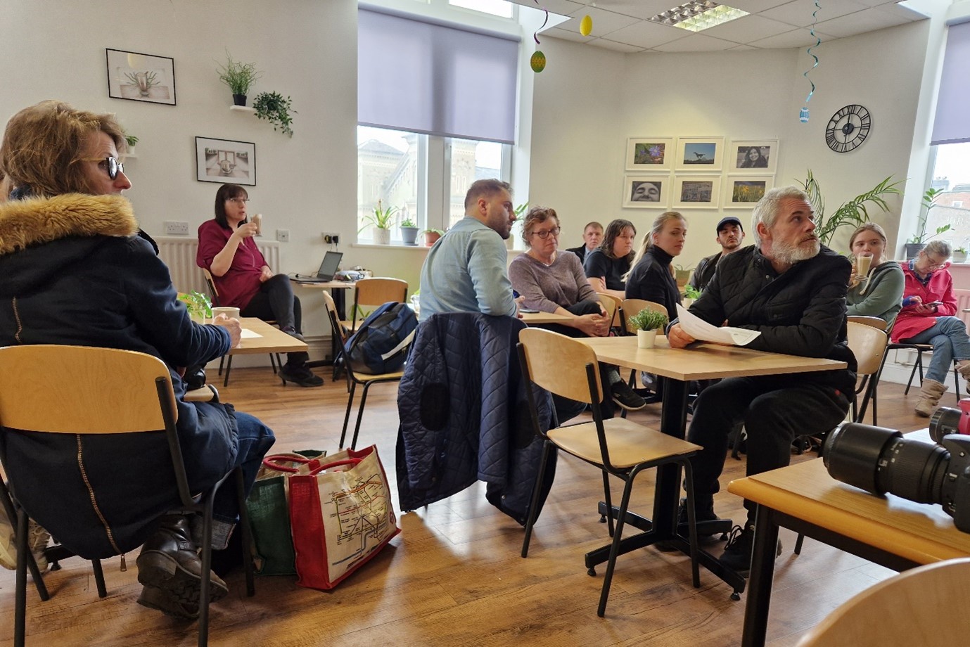 A room of people sat around multiple tables, looking at a point off camera.