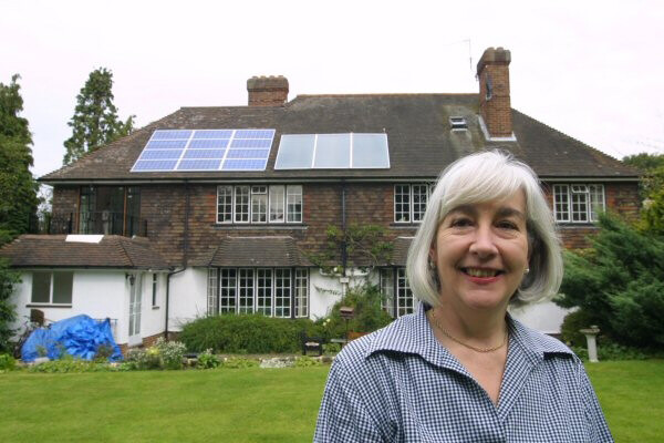 Photograph of a smiling woman's head and shoulders. Behind her is a house with solar panels on the roof.
