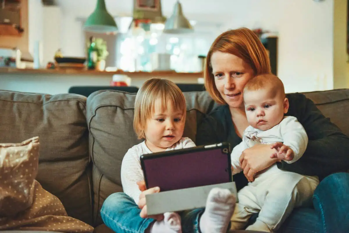 a woman sits on the sofa holding two babies, all looking at a tablet.