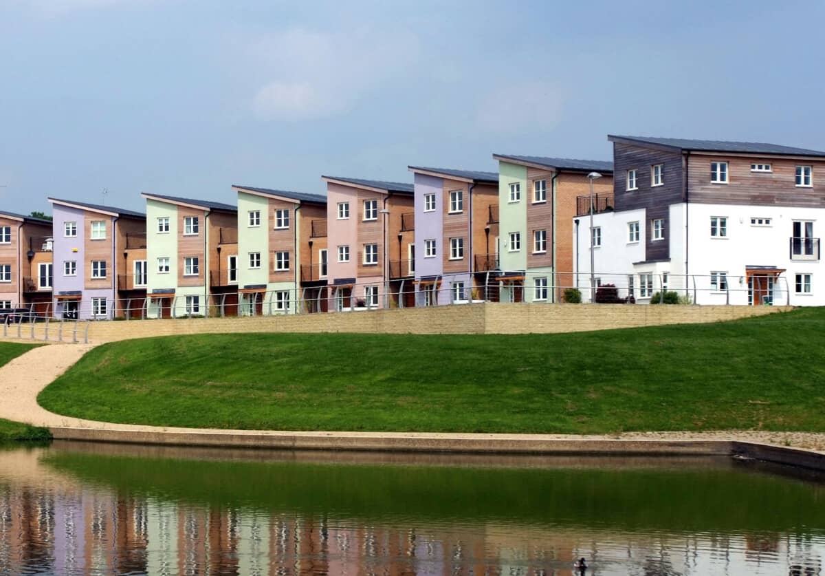 A row of newly built houses. They are reflected in a lake in the foreground.