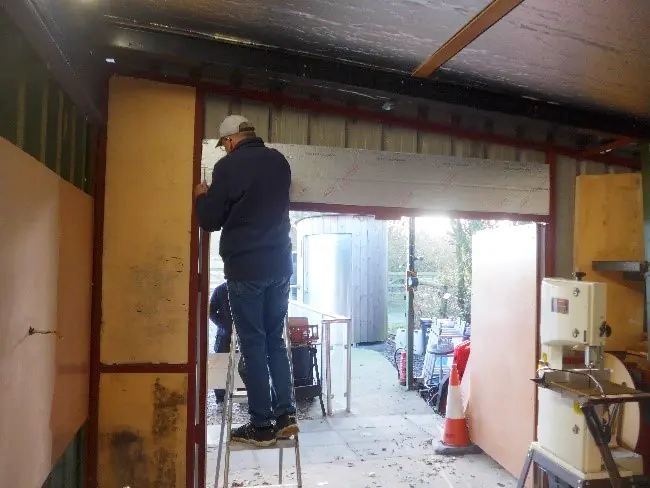 A man standing on a ladder inside a shed, fixing internal wall insulation above an open door.