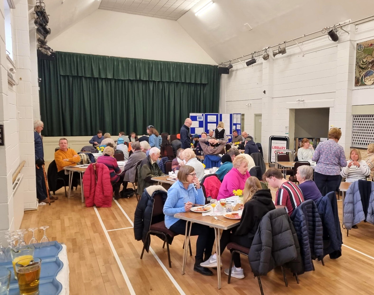 Community hall interior with many people sitting around tables, eating and talking.