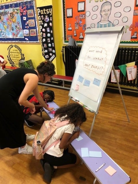 A woman and two children learn over a desk, looking at colourful post it notes. A whiteboard above them reads "What have you learnt about energy?"