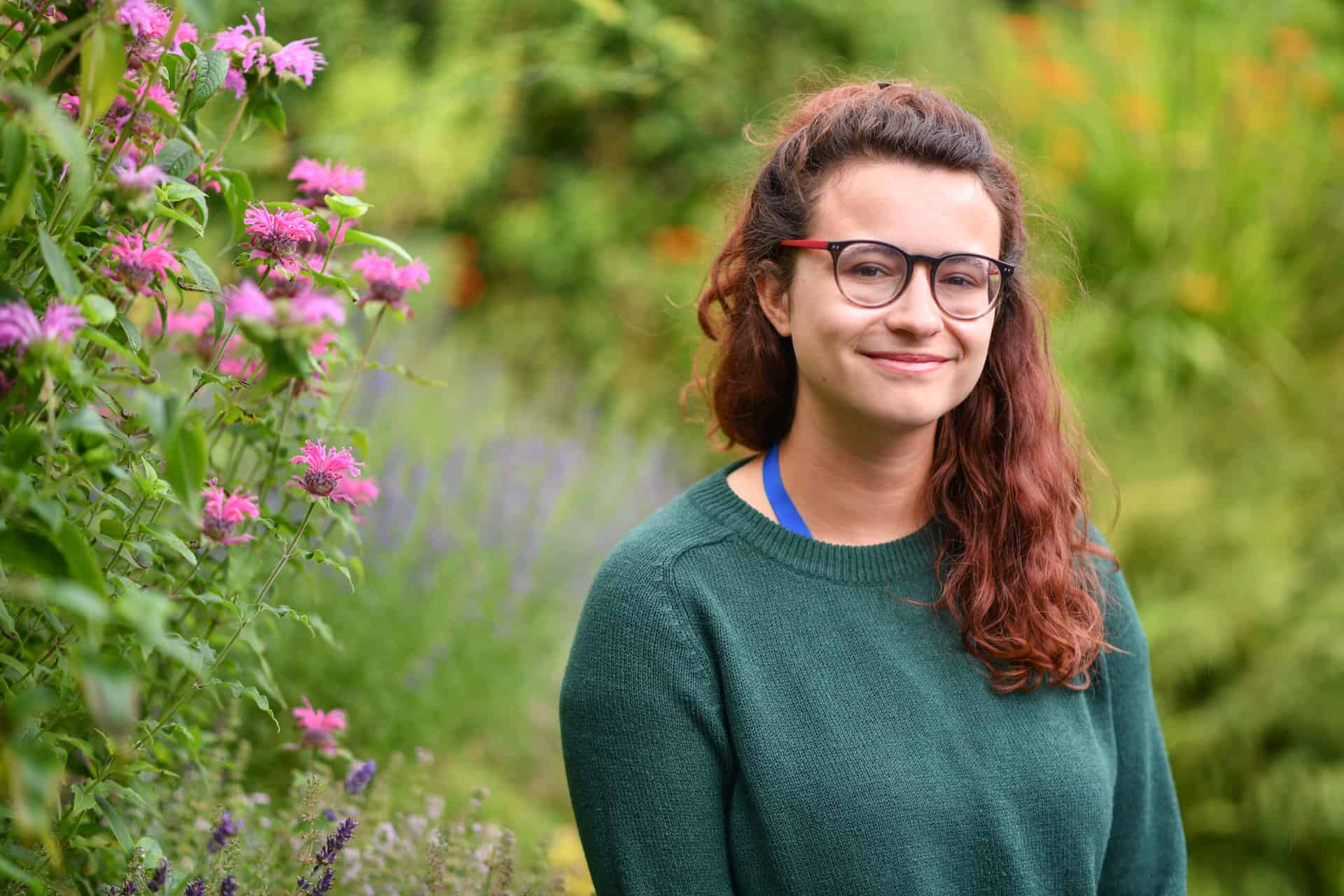 A woman with dark hair and glasses standing outside, surrounded by green plants and trees, smiling and looking at the camera.