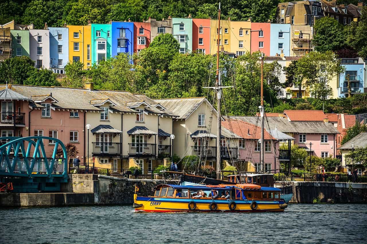Ferry on a river, with a row of houses painted bright colours in the background.
