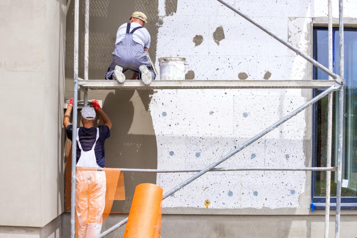 Two workers install solid wall insulation to the exterior of a building. They are spreading mortar onto polystyrene blocks.