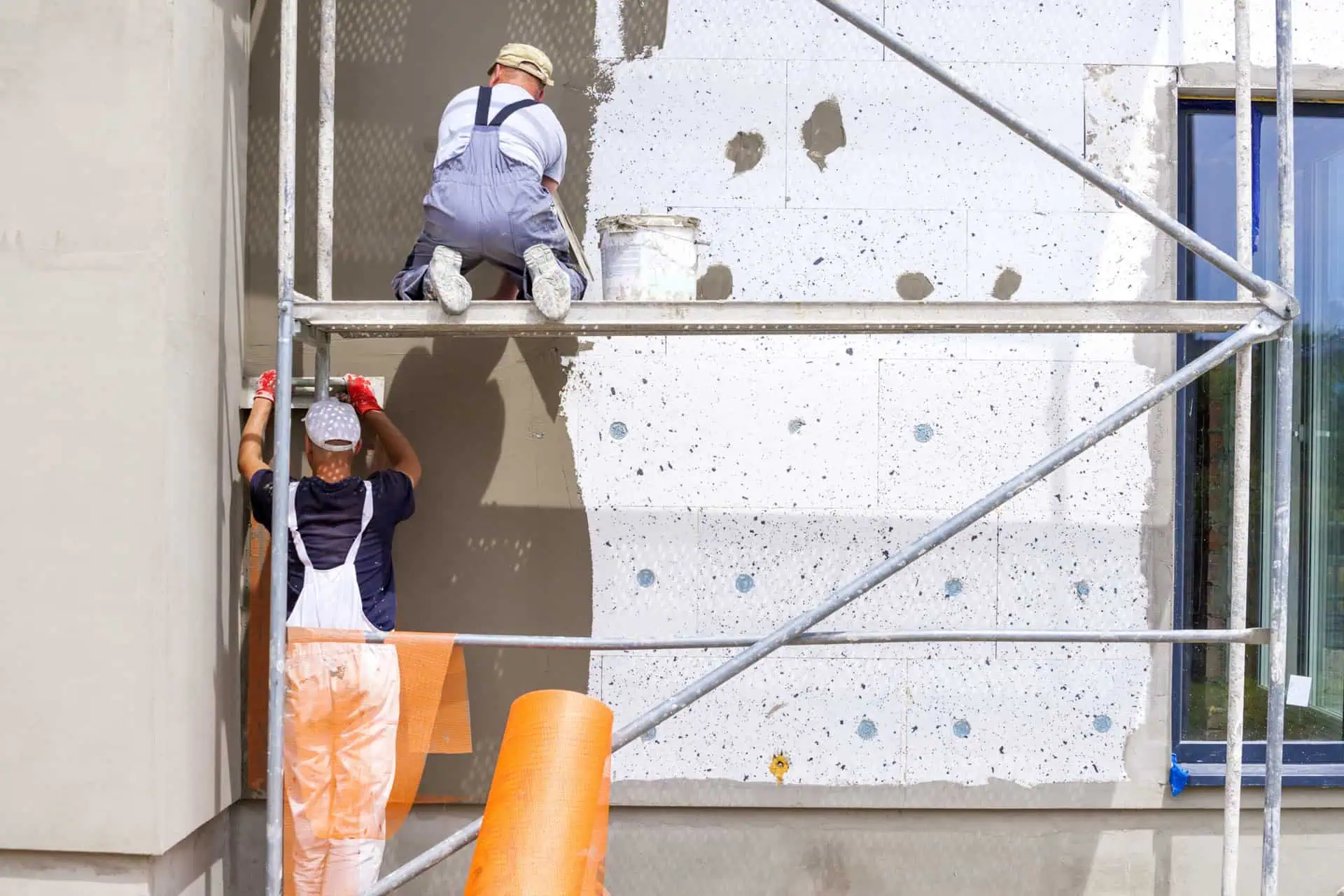 Two workers install solid wall insulation to the exterior of a building. They are spreading mortar onto polystyrene blocks.