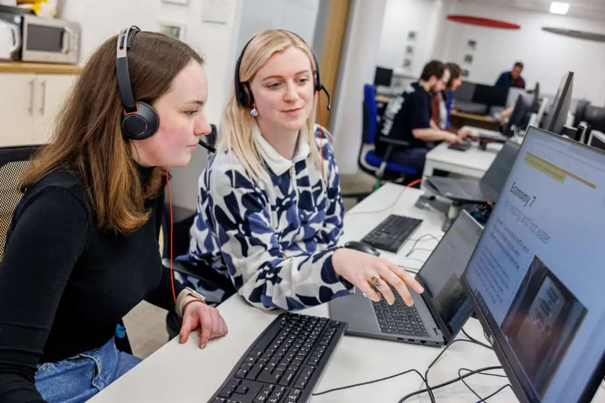 Two women wearing headsets sit at desks, looking at a screen showing information about Economy 7 tariffs.