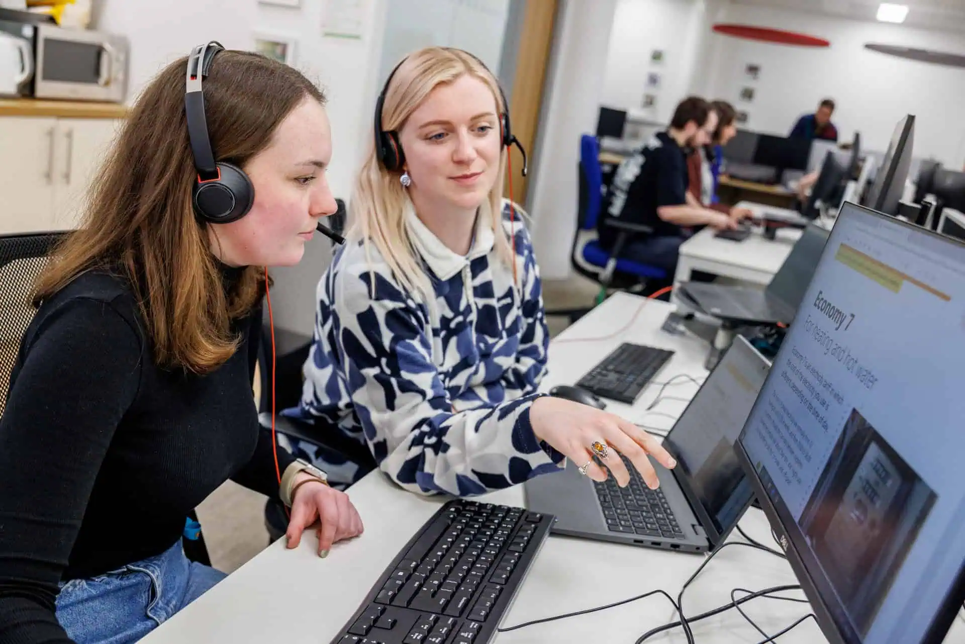 Two women wearing headsets sit at desks, looking at a screen showing information about Economy 7 tariffs.