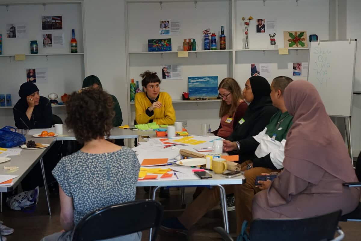 A diverse group of people sat around a table during a workshop. On the table are post it notes and pieces of paper.