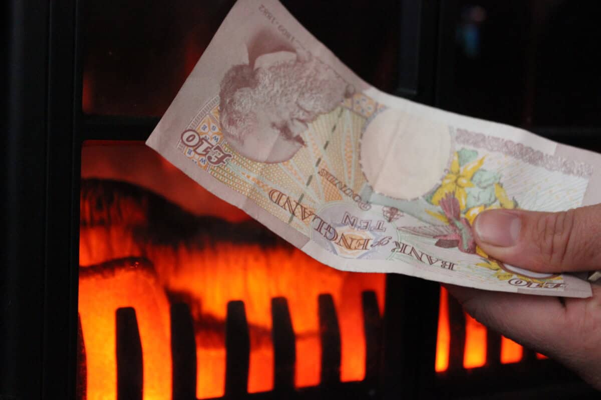 A hand holds a ten pound note in front of a burning fire