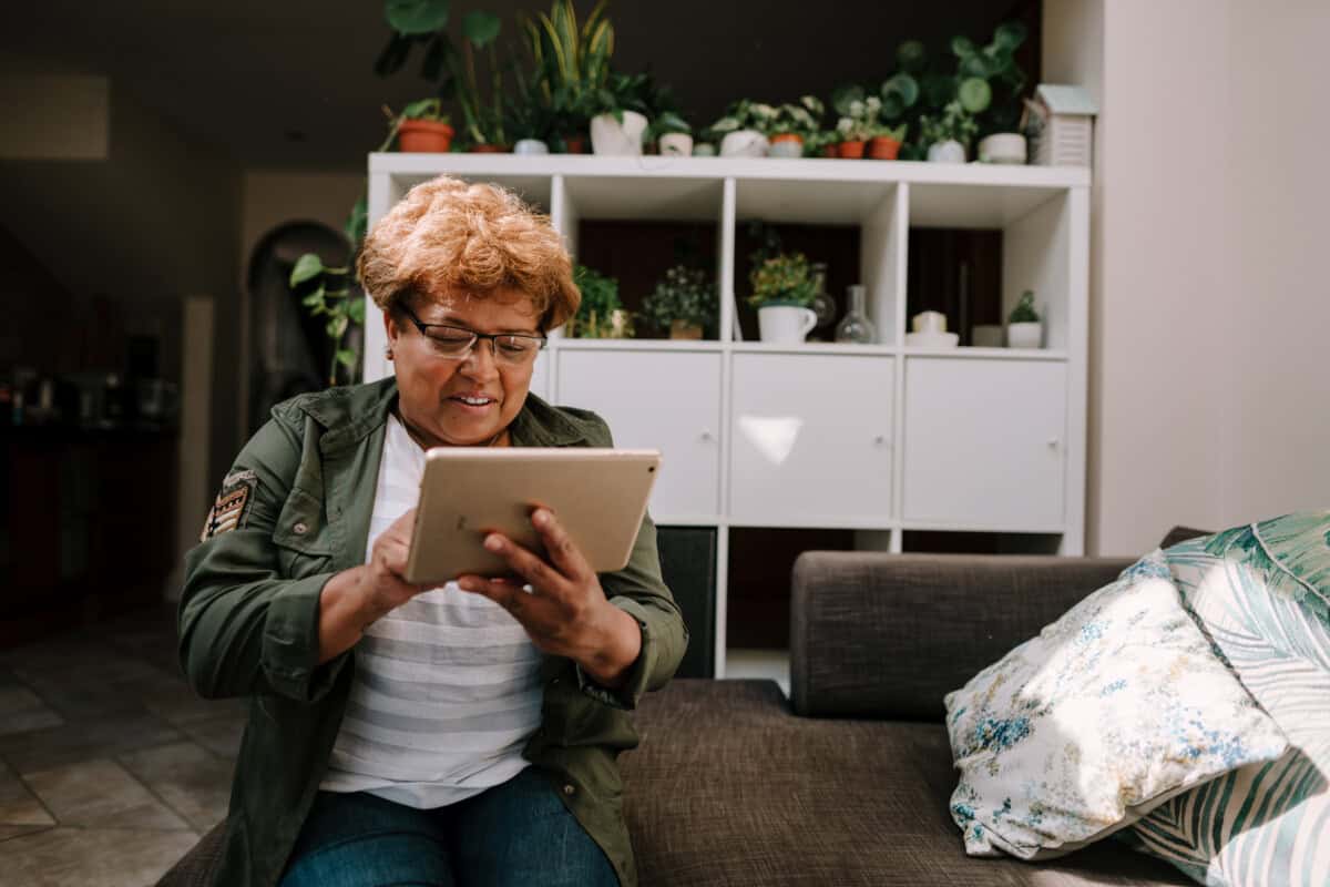 Older woman sitting on a sofa, looking down at a tablet she holds with both hands. She is smiling.