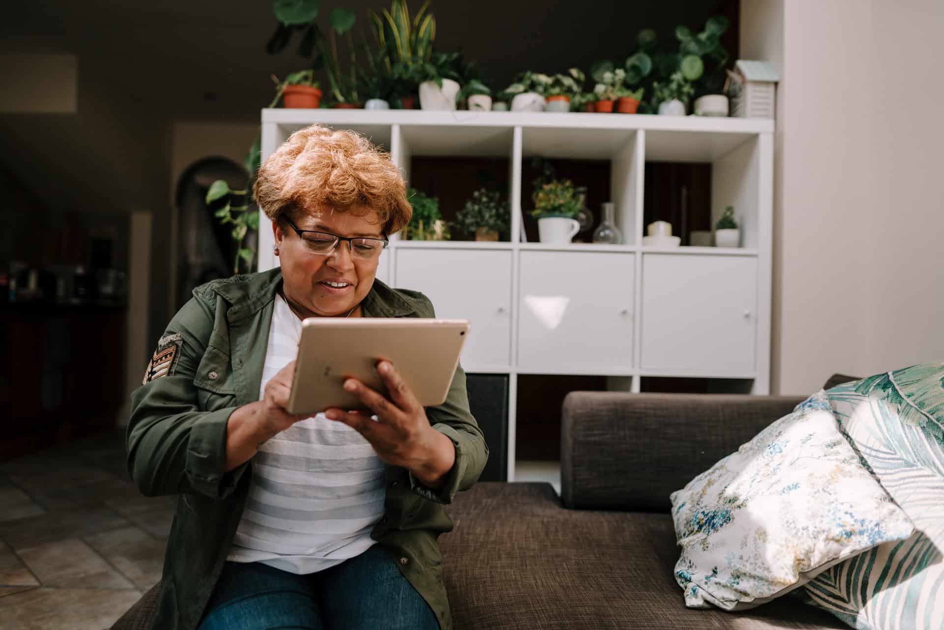 Older woman sitting on a sofa, looking down at a tablet she holds with both hands. She is smiling.