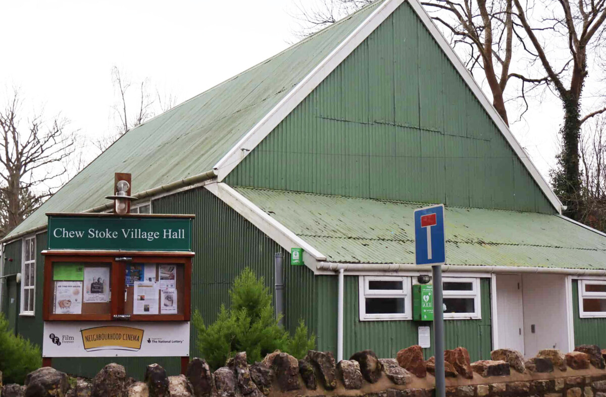 The outside of Chew Stoke Village Hall - a green corrugated building with a noticeboard outside