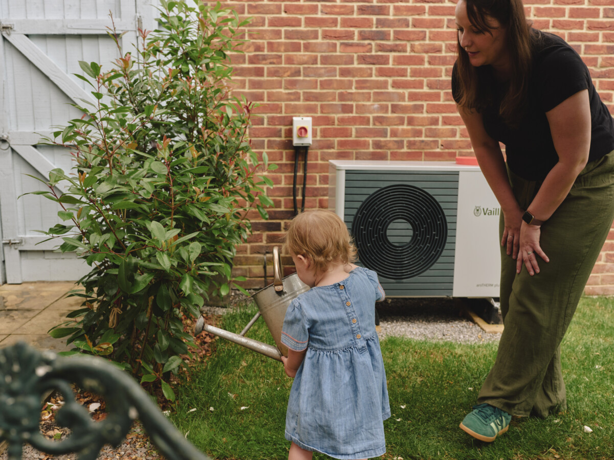 A mother and her child watering plants in the garden with a heat pump on the brick wall behind them