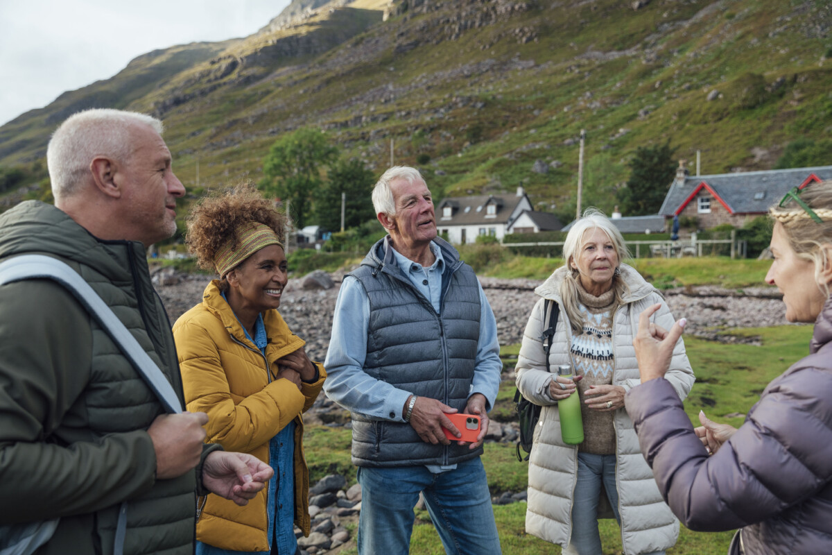 A shot of a small group of adults standing on the shore of a sea loch in Torridon, Scotland. They are all wearing warm clothing and looking at one of the group members who is talking. The shoreline is rocky and there are a couple of houses and rugged hills behind them.