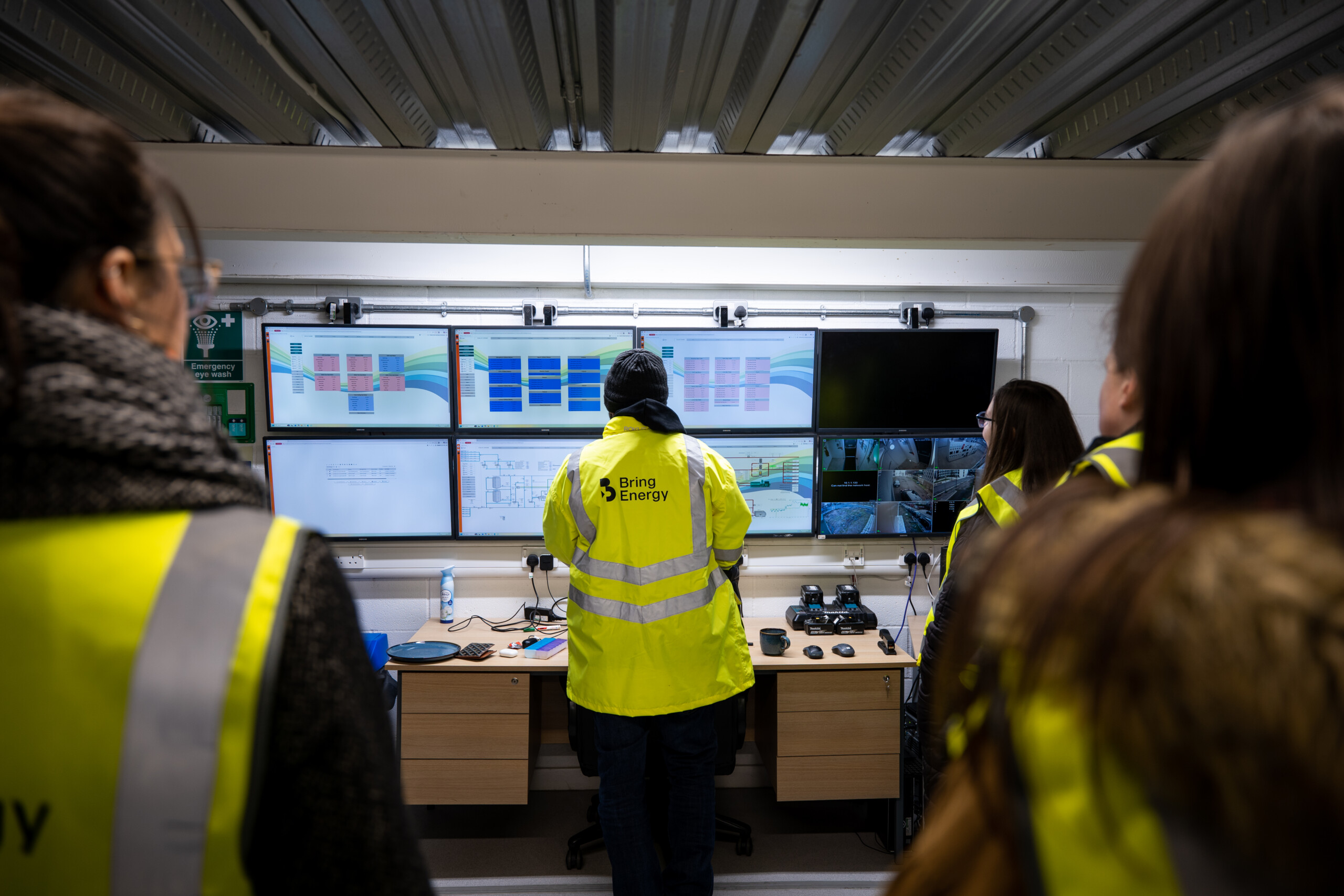 3 people in high vis jackets in front of a wall of monitors showing data , presumably at a power generation facility