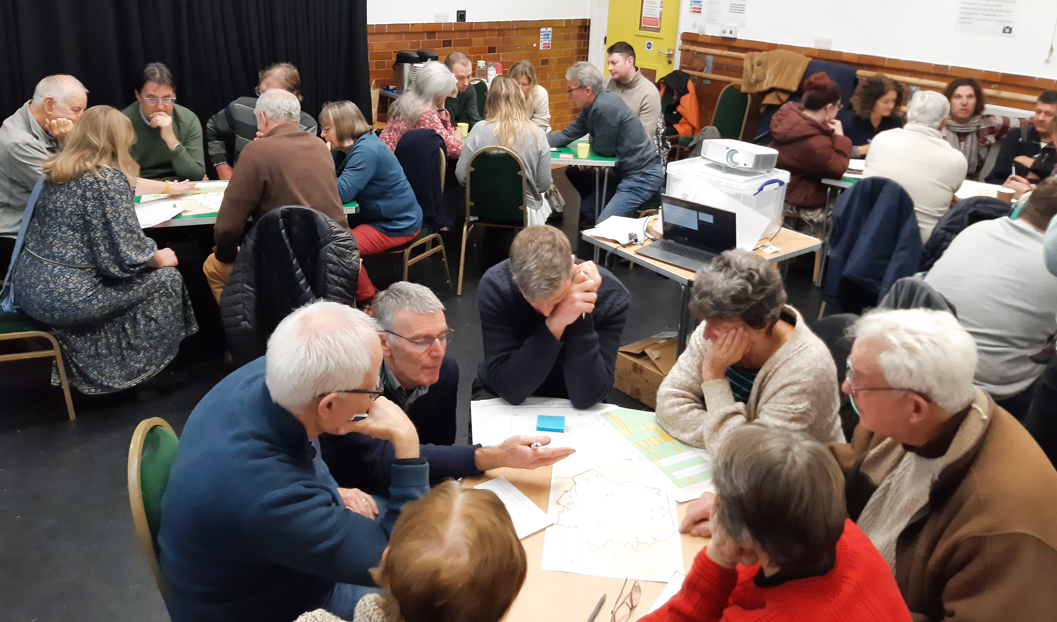 Around 30 people sit around several tables in large room in a village hall or similar building engaged in discussion.