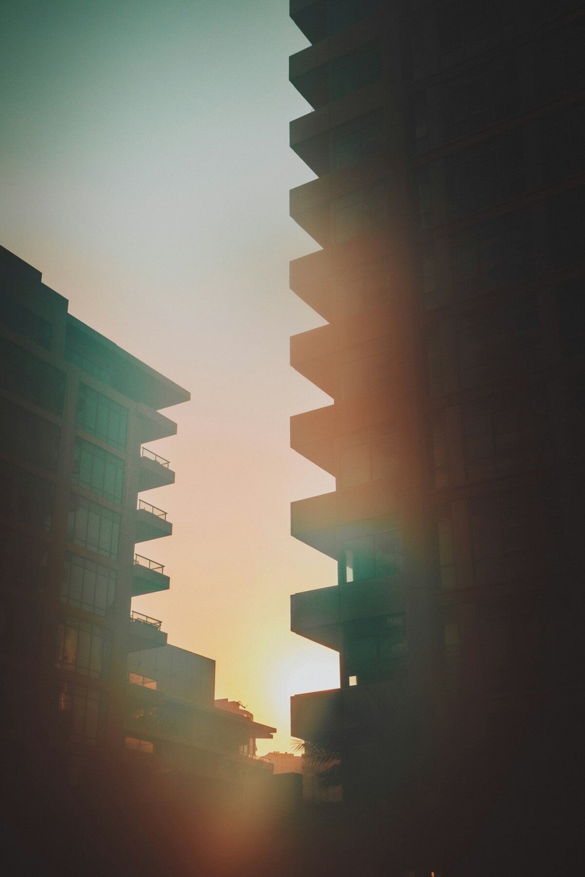 Low-angle view looking up between two modern residential tower blocks in hazy heat, with a glowing orange and yellow sunset visible between them, conveying the intensity of urban heat in a built environment.
