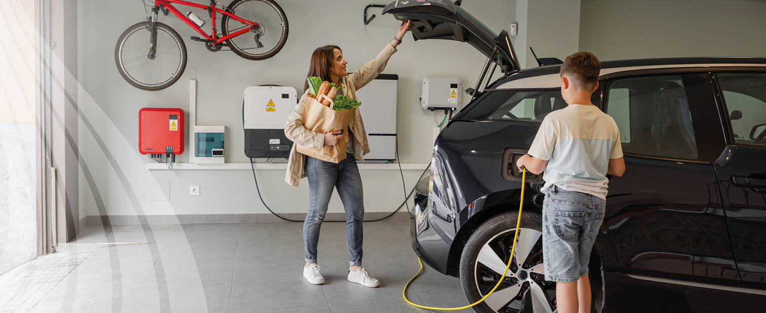 A woman takes groceries from the boot of her electric car which is parked in a spacious new garage while her son recharges it. In the background are a solar converter and domestic battery.